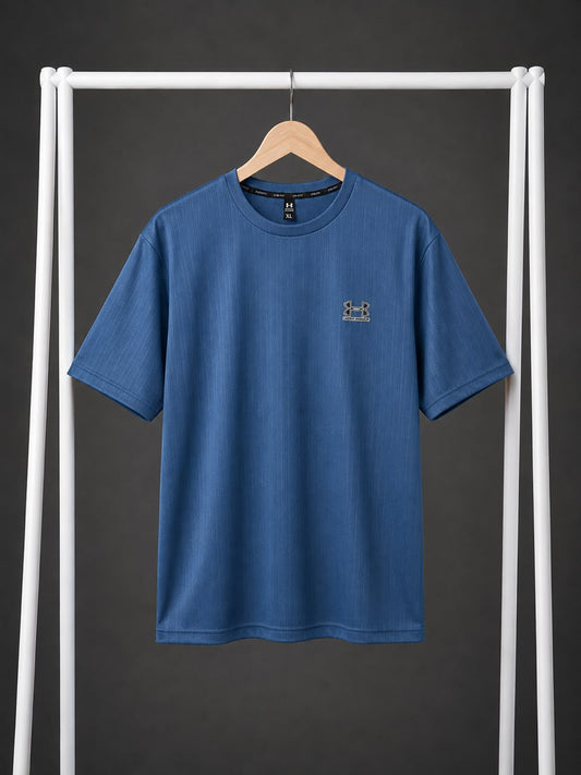 A blue short-sleeve crewneck t-shirt with a small embroidered Under Armour logo on the left chest, hanging on a wooden hanger from a white clothing rack against a dark gray background.