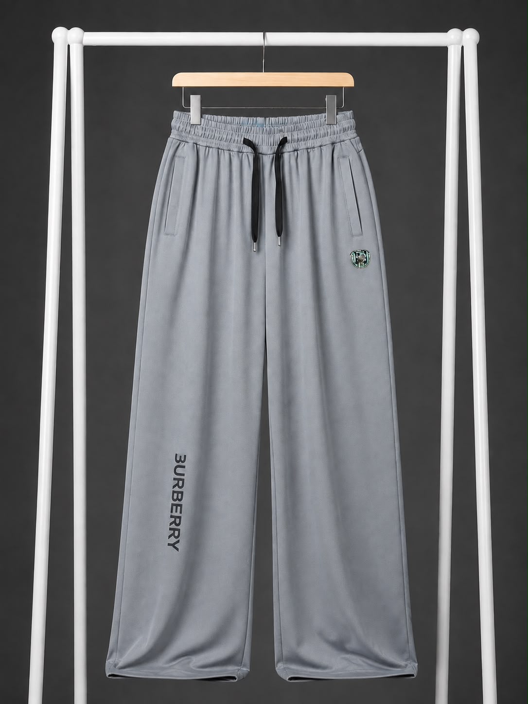 Light gray wide-leg sweatpants with a black drawstring waist, featuring 'BURBERRY' printed vertically on one leg and a small embroidered crest on the other pocket area, hanging on a wooden hanger against a dark gray background.