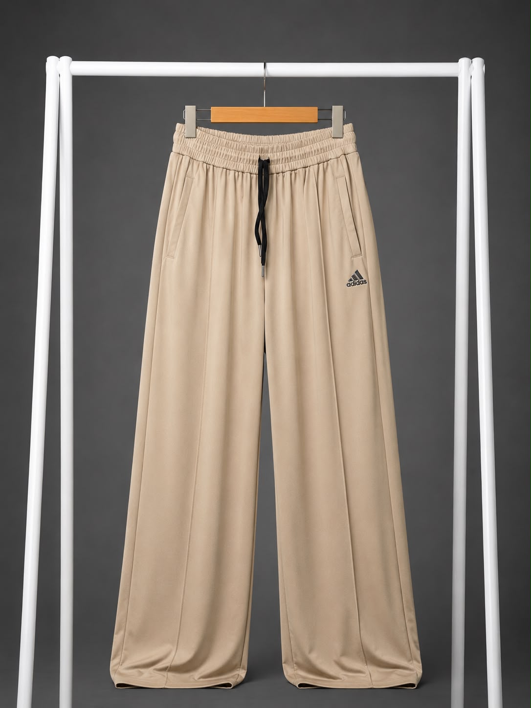 Beige wide-leg athletic pants with an elastic waistband, black drawstring, and a small Adidas logo on the left thigh, hanging on a white clothing rack against a dark gray background.