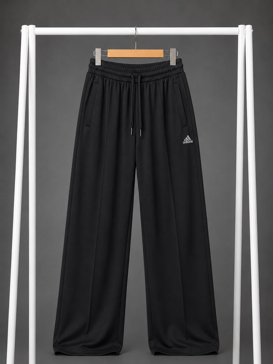 Black wide-leg athletic pants with an elastic drawstring waist and a small white Adidas logo on the left thigh, hanging on a wooden hanger from a white clothing rack against a dark gray background.