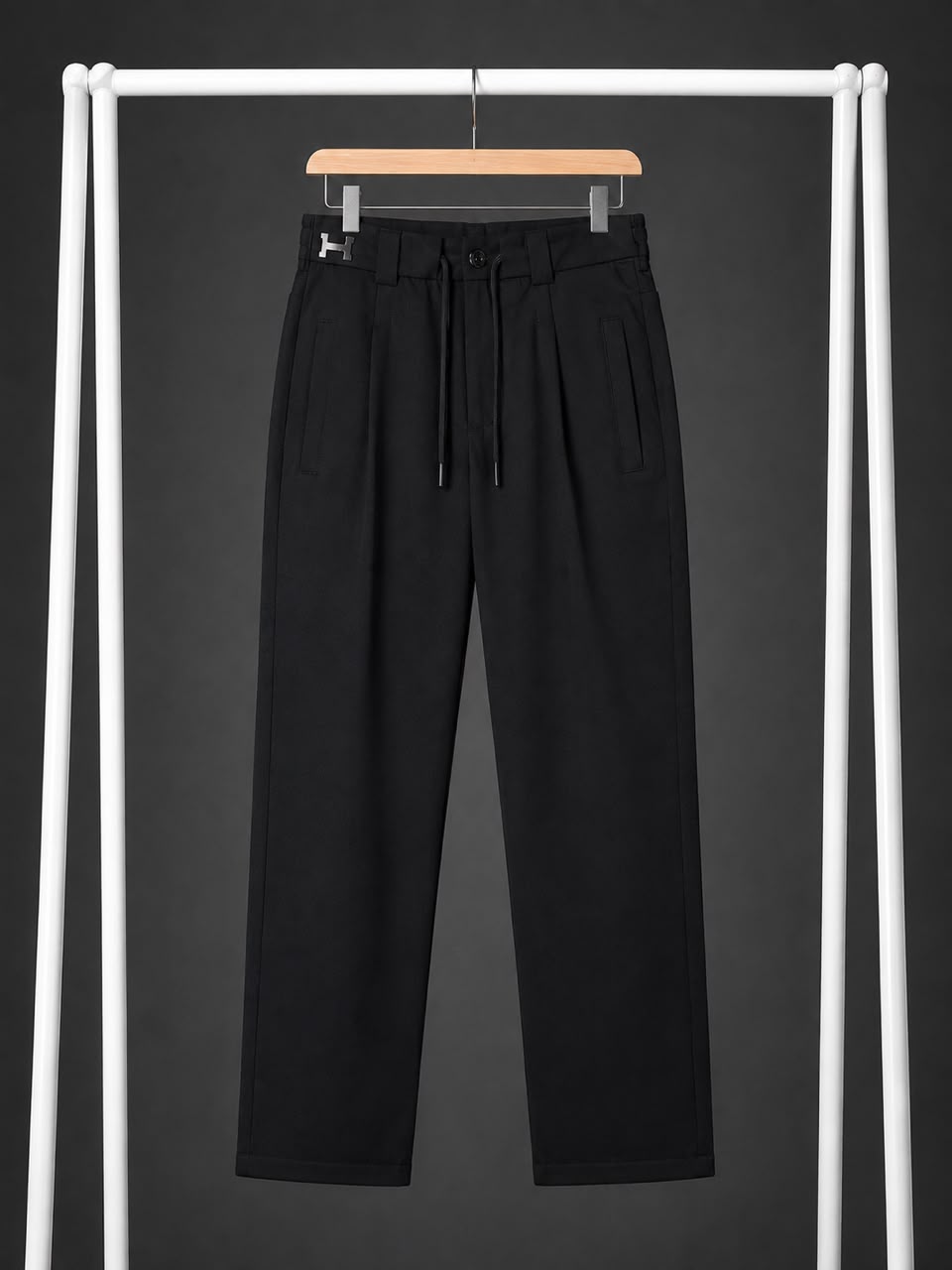 Black high-waisted trousers with a drawstring waist and side pockets, hanging on a wooden hanger from a white clothing rack against a dark gray background.