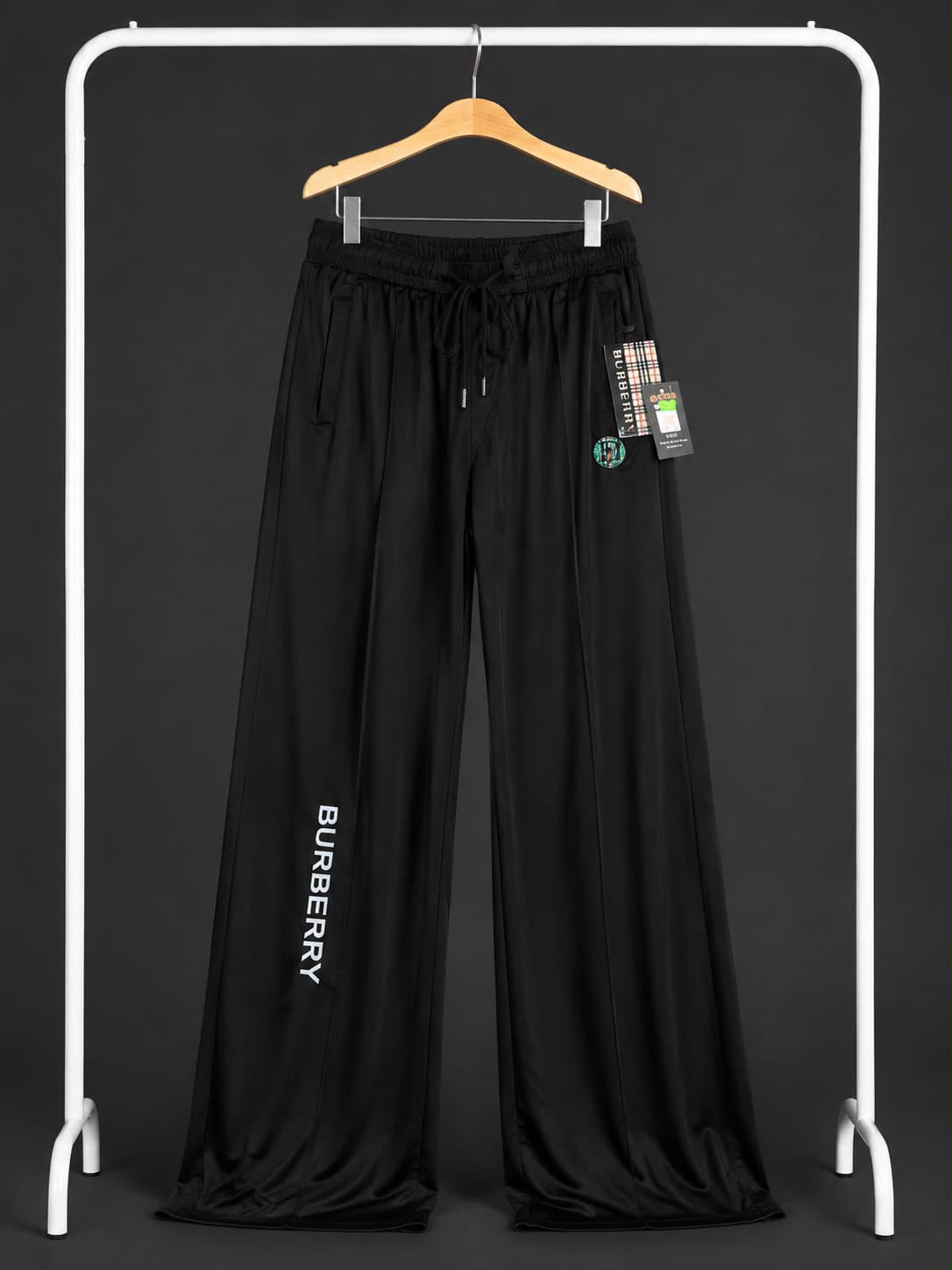 Black wide-leg sweatpants with a drawstring waist and vertical BURBERRY text on one leg, hanging on a wooden hanger from a white clothing rack against a dark background.