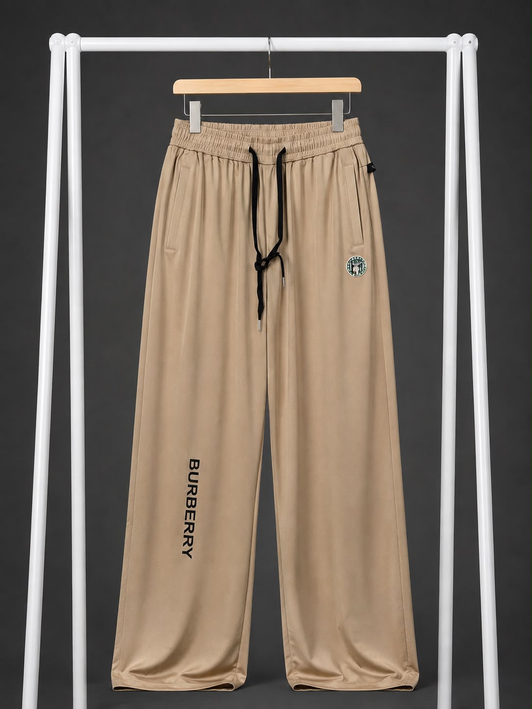 A pair of Burberry beige wide-leg sweatpants hanging on a wooden hanger from a white clothing rack. The pants feature a black drawstring waist, a black vertical BURBERRY logo printed on the left leg, and a small green and white embroidered logo on the right hip.