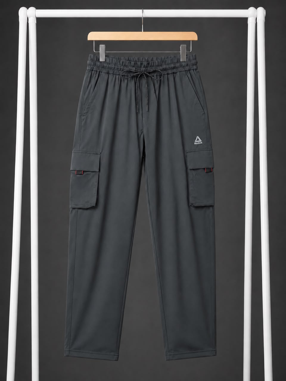 Dark gray cargo pants with an elastic drawstring waist and side cargo pockets, hanging on a wooden hanger against a dark gray background framed by white vertical bars.
