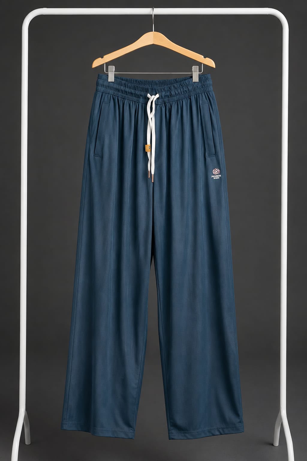 A pair of wide-leg, relaxed-fit pants in a dark slate blue color, hanging on a wooden hanger from a white clothing rack. The pants feature an elastic waistband with a white drawstring, side pockets, and a small embroidered logo on the left thigh.