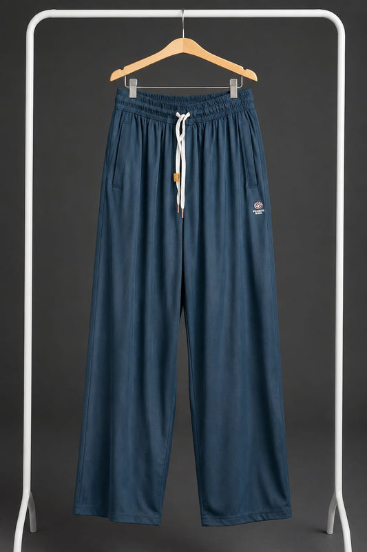 A pair of wide-leg, relaxed-fit pants in a dark slate blue color, hanging on a wooden hanger from a white clothing rack. The pants feature an elastic waistband with a white drawstring, side pockets, and a small embroidered logo on the left thigh.