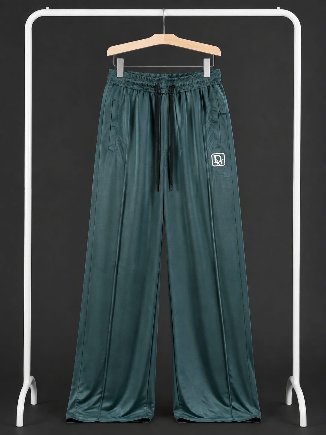 A pair of wide-leg, dark teal-green pants hanging on a wooden hanger from a white clothing rack against a dark gray background. The pants feature an elastic waistband with a drawstring and a small white square logo patch on the left thigh.