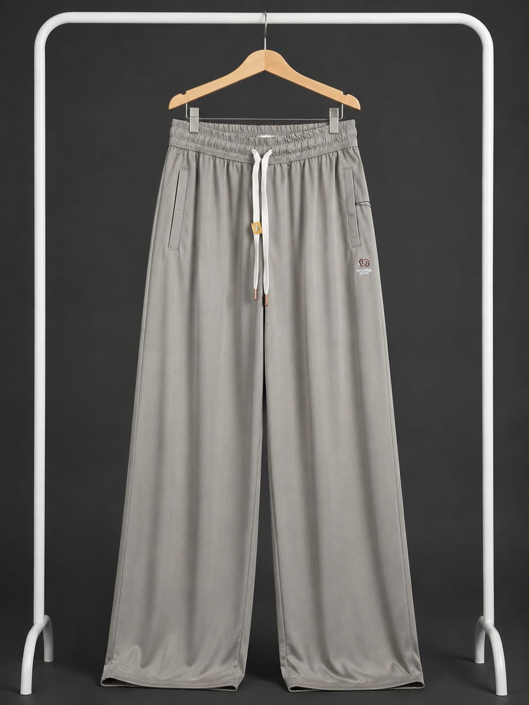 Light gray wide-leg sweatpants with an elastic waistband, white drawstring, and side pockets, hanging on a wooden hanger from a white clothing rack against a dark gray background.