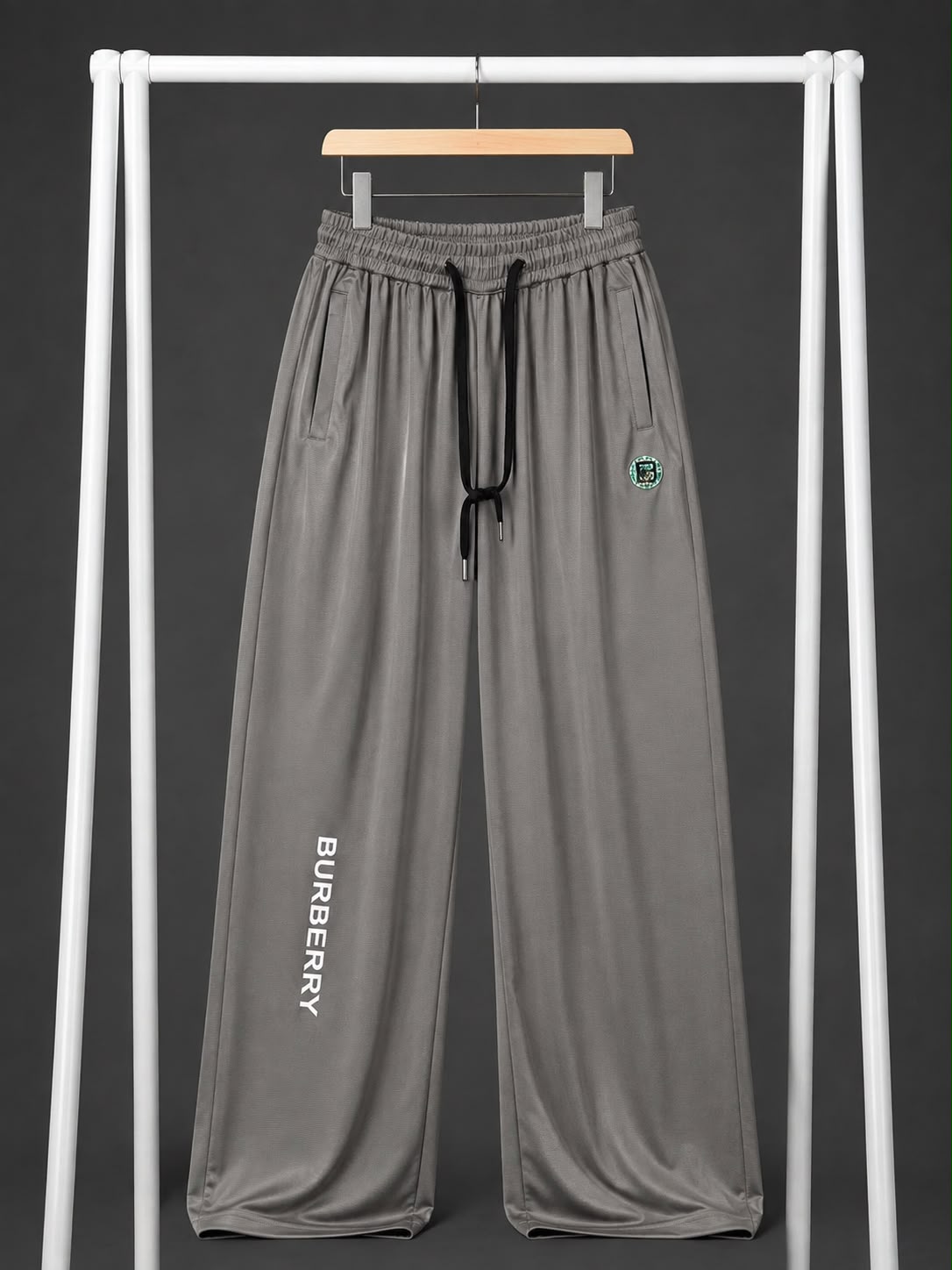 Gray wide-leg sweatpants with a black drawstring, featuring a vertical 'BURBERRY' logo printed on one leg and a small circular green crest patch on the other thigh, hanging on a wooden hanger from a white clothing rack against a dark gray background.