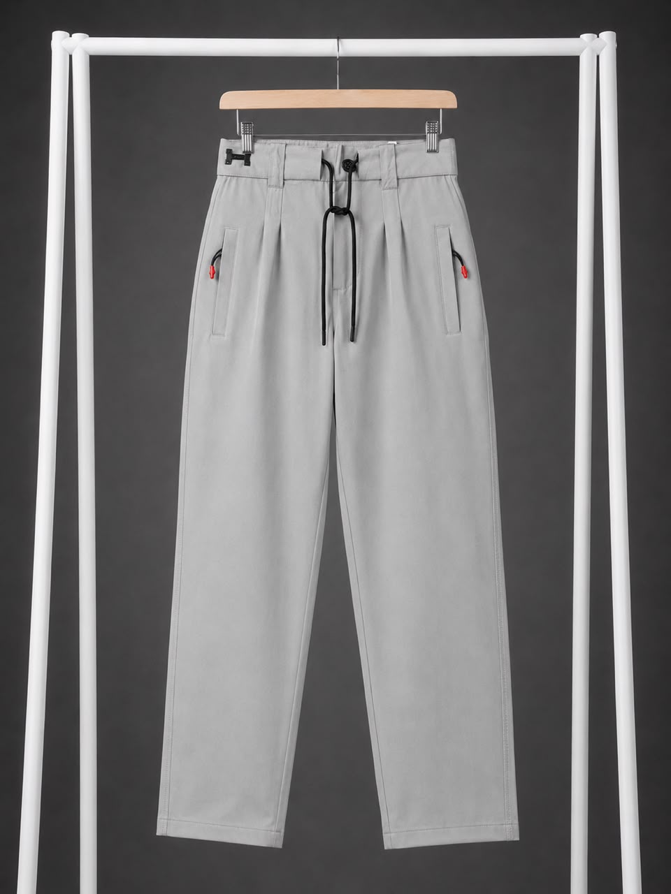 Light gray wide-leg trousers with pleats, a black drawstring waist, and small red embroidered details on the side pockets, hanging on a wooden hanger from a white clothing rack against a dark gray background.