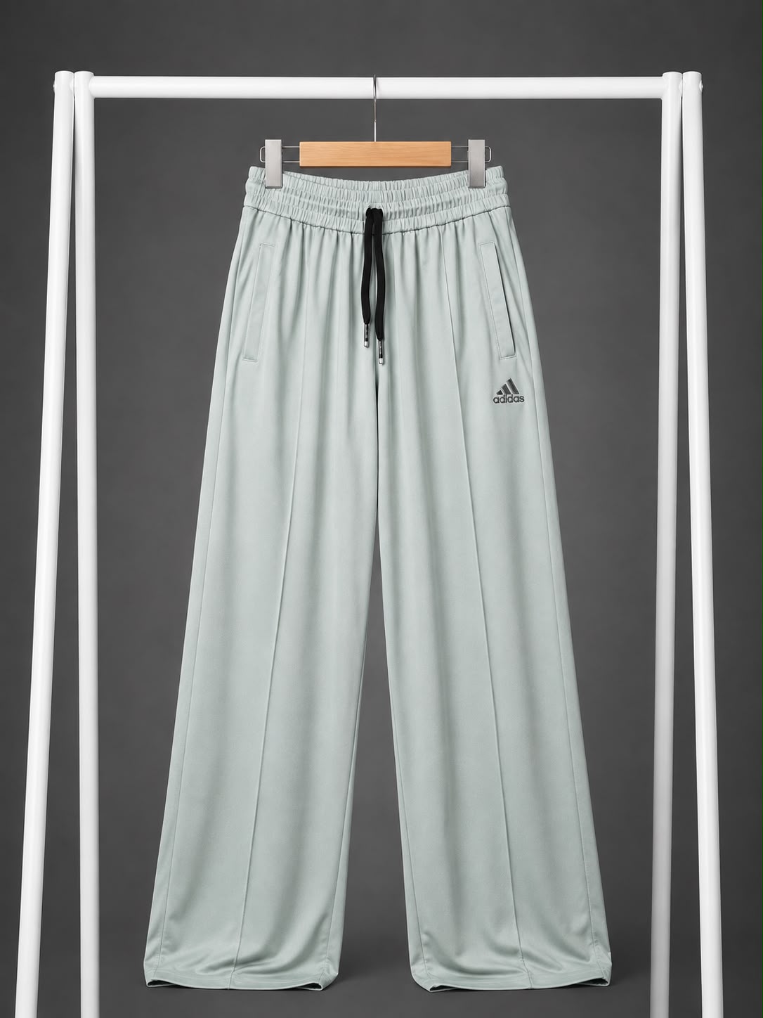 A pair of light sage green wide-leg athletic pants hanging from a wooden hanger on a white clothing rack against a dark gray background. The pants feature an elastic waistband with a black drawstring and a small black Adidas logo on the left thigh.