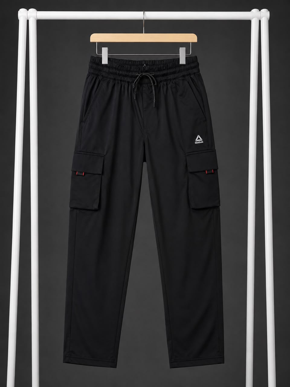 Black cargo pants with an elastic drawstring waist and side cargo pockets, hanging on a wooden hanger from a white clothing rack against a dark gray background.