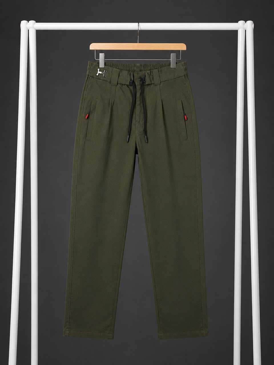 Olive green drawstring trousers hanging on a wooden hanger from a white clothing rack against a dark gray background.