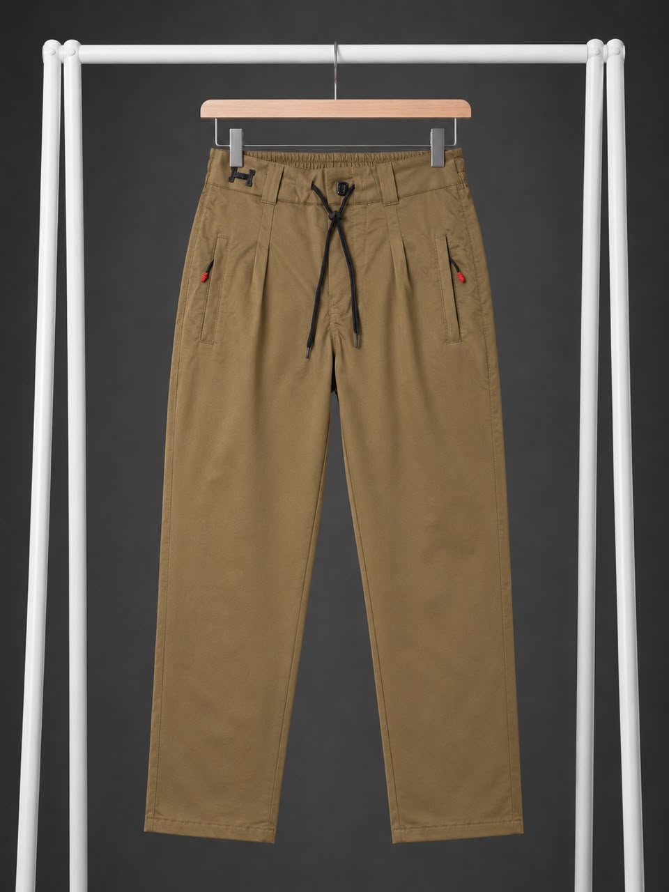 A pair of men's relaxed-fit trousers in a warm khaki-brown color, hanging on a wooden hanger from a white clothing rack against a dark gray background. The pants feature a black drawstring waist, pleated front, and small red accents on the side pockets.