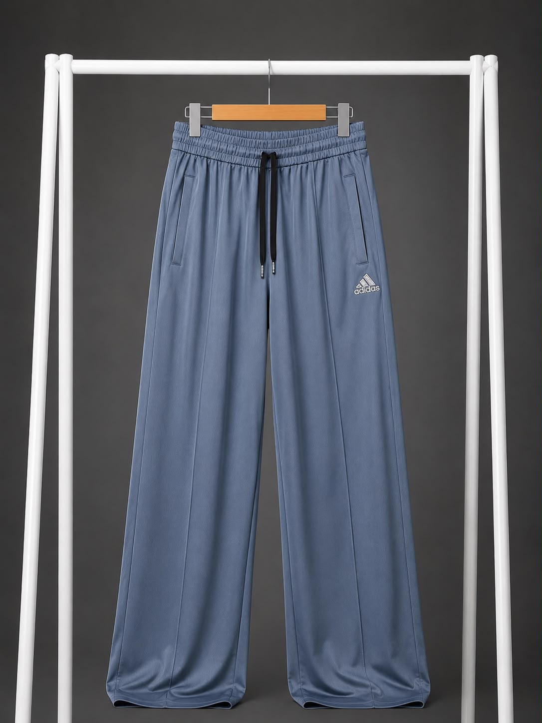 A pair of wide-leg athletic pants in dusty blue, hanging on a wooden hanger from a white clothing rack. The pants feature an elastic waistband with a black drawstring and a small white Adidas logo on the left thigh.