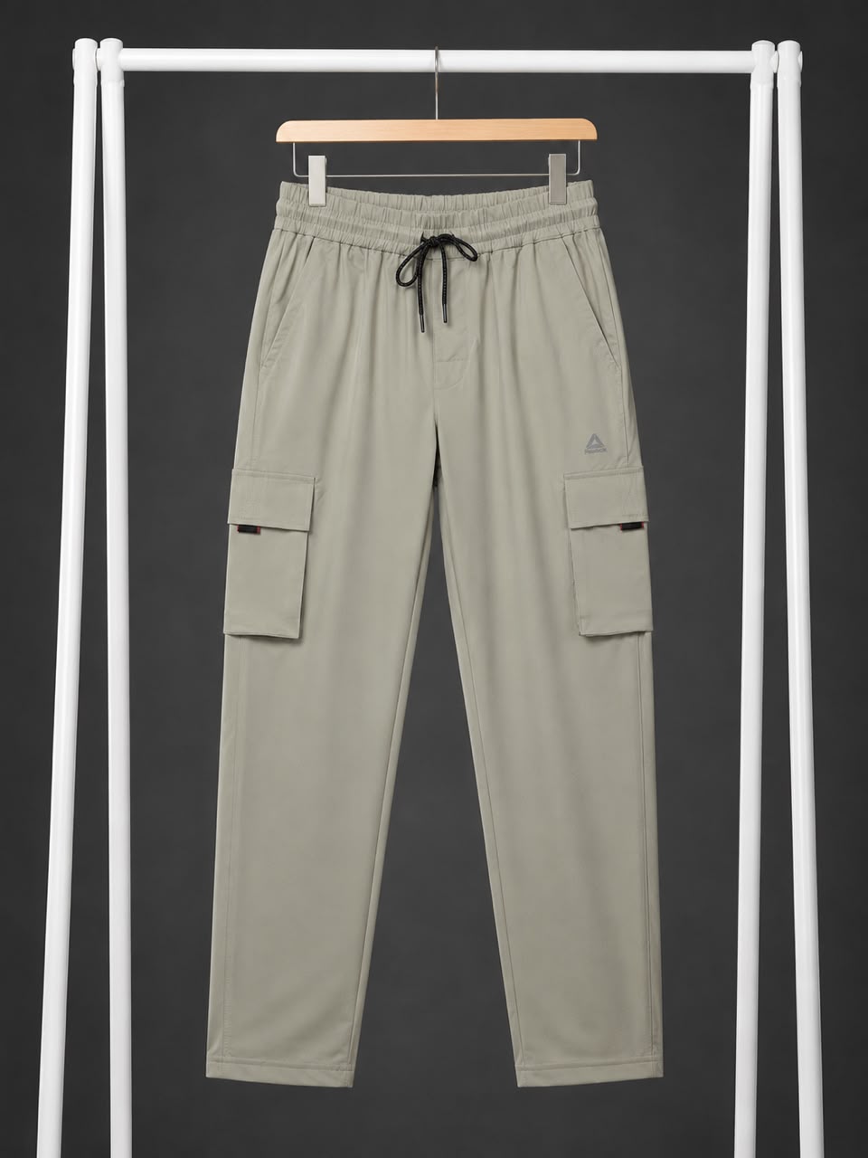 A pair of light khaki cargo pants hanging on a wooden hanger from a white clothing rack, featuring side cargo pockets and a drawstring waist.
