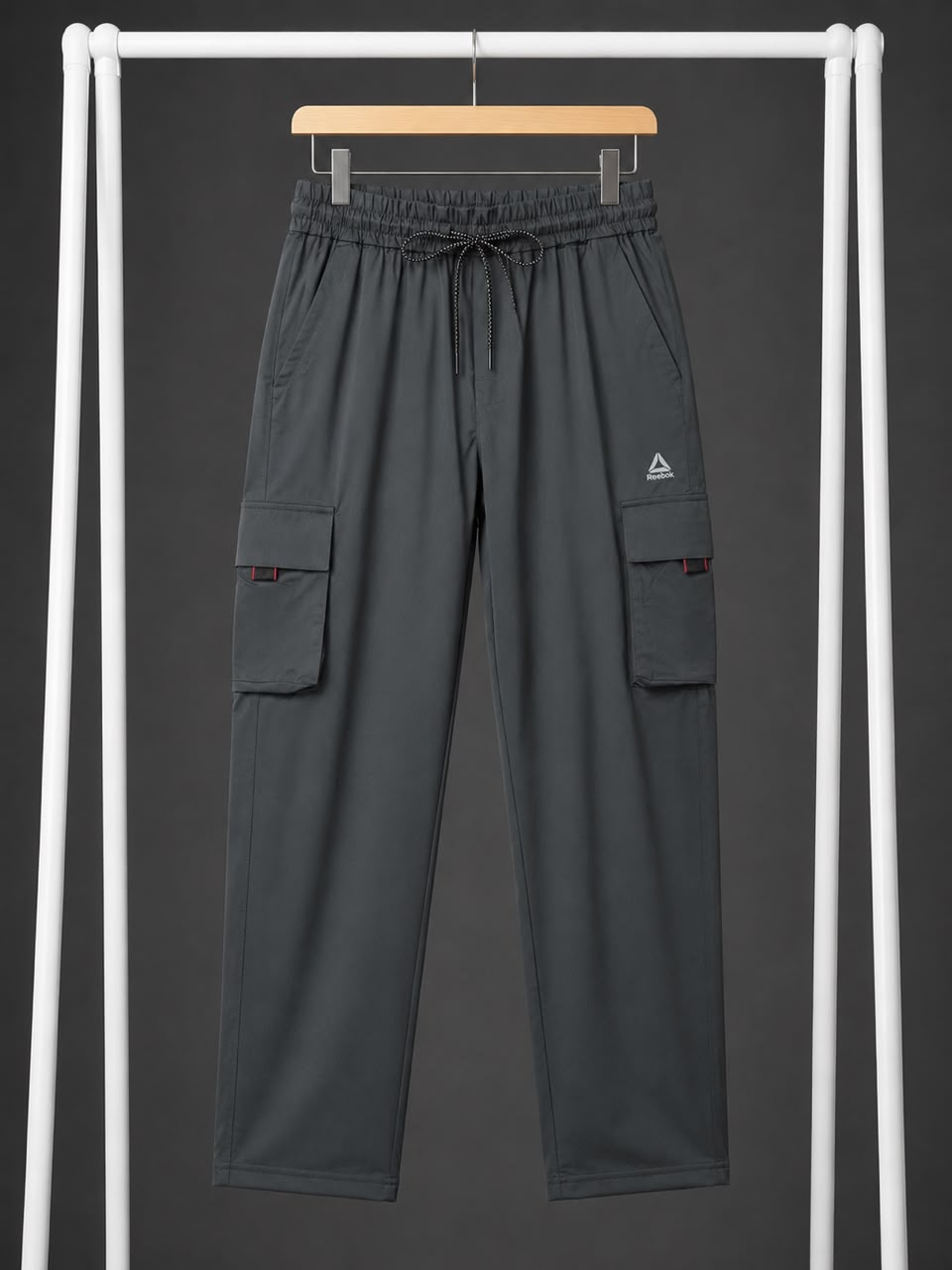 Dark gray cargo pants with an elastic drawstring waist and side cargo pockets, hanging on a wooden hanger against a dark gray background framed by white vertical bars.