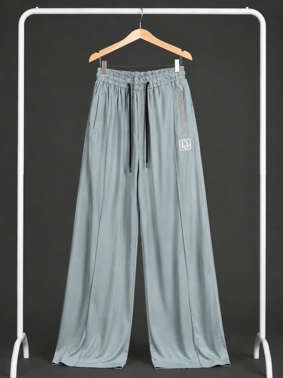 A pair of wide-leg lounge pants in a muted sage green, hanging on a wooden hanger from a white clothing rack against a dark gray background.