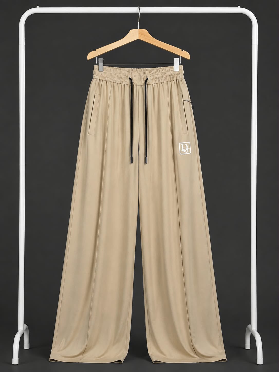 A pair of wide-leg beige pants hanging on a wooden hanger from a white clothing rack, featuring an elastic waistband with black drawstrings and a small white square logo patch on the left thigh.