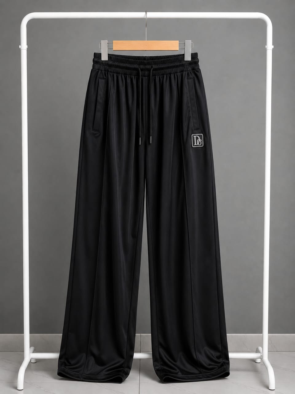 Black wide-leg sweatpants with an elastic drawstring waist and a small white embroidered logo on the left thigh, displayed on a white clothing rack against a gray background.