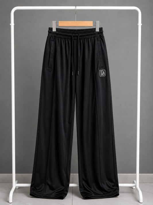 Black wide-leg sweatpants with an elastic drawstring waist and a small white embroidered logo on the left thigh, displayed on a white clothing rack against a gray background.