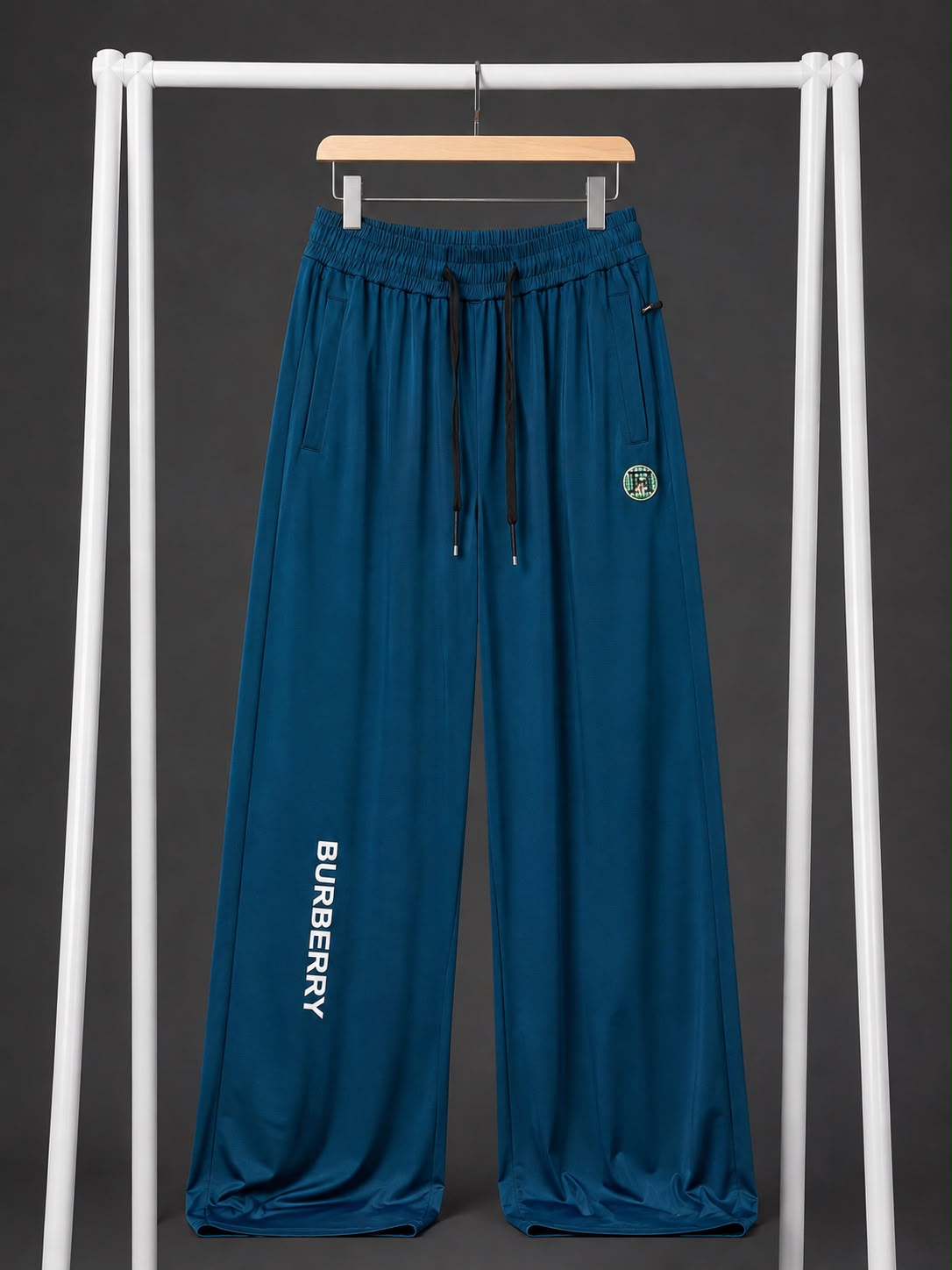 Teal blue wide-leg sweatpants with a black drawstring, featuring 'BURBERRY' printed vertically in white on the left leg and a circular Burberry logo patch on the right hip, hanging on a wooden hanger from a white clothing rack against a dark gray background.