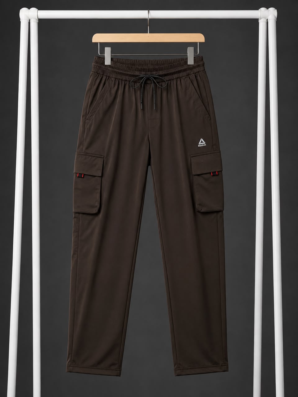 Dark brown cargo pants with an elastic drawstring waist and side cargo pockets, hanging on a wooden hanger from a white clothing rack against a dark gray background.