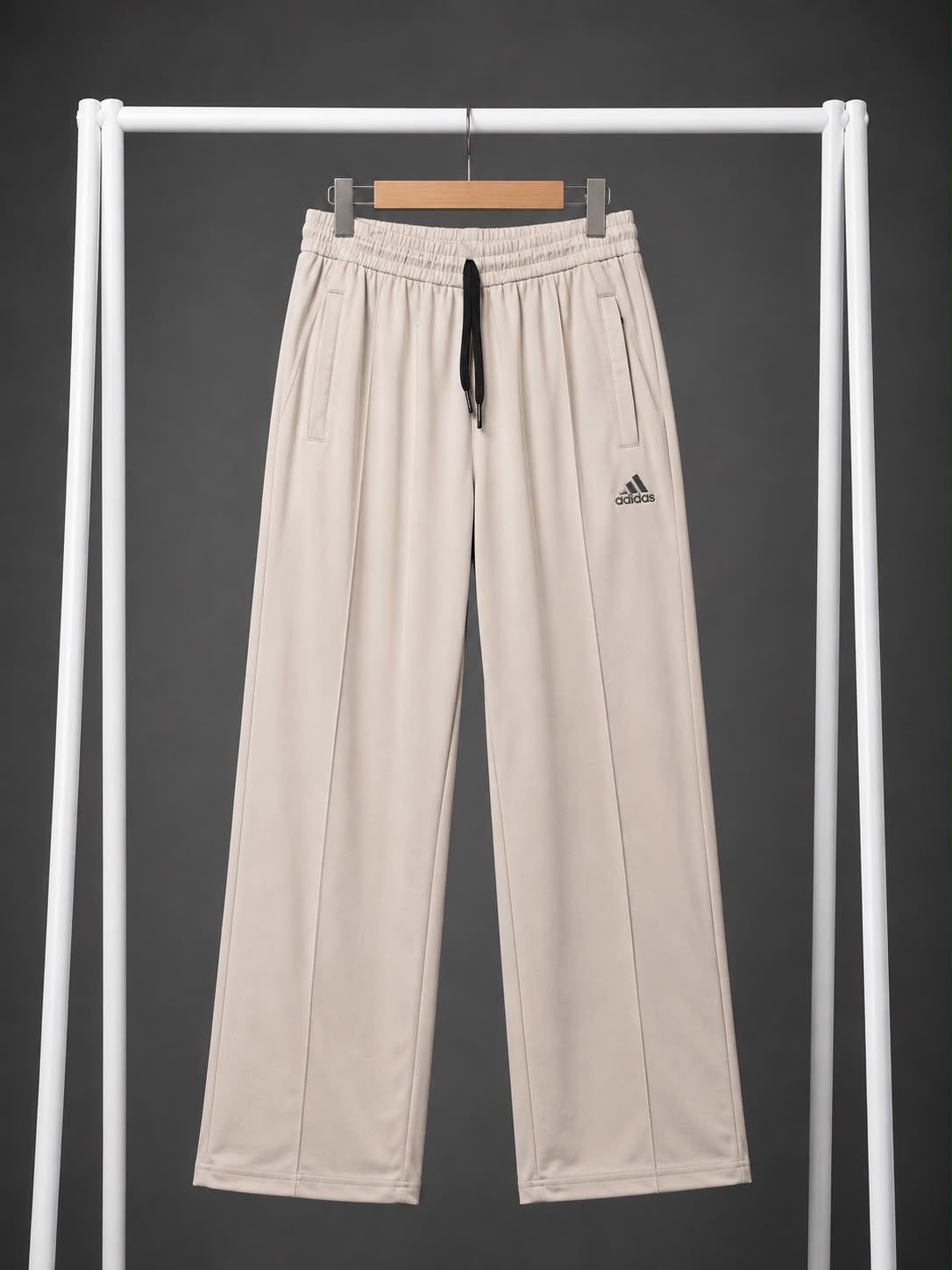 Beige wide-leg sweatpants with an elastic waistband, black drawstring, and a small black Adidas logo on the left thigh, hanging on a white clothing rack against a dark gray background.
