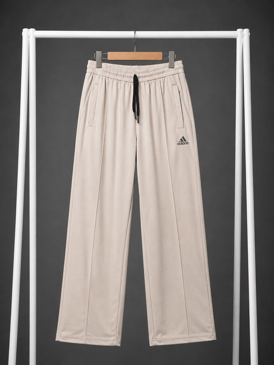 Beige wide-leg sweatpants with an elastic waistband, black drawstring, and a small black Adidas logo on the left thigh, hanging on a white clothing rack against a dark gray background.