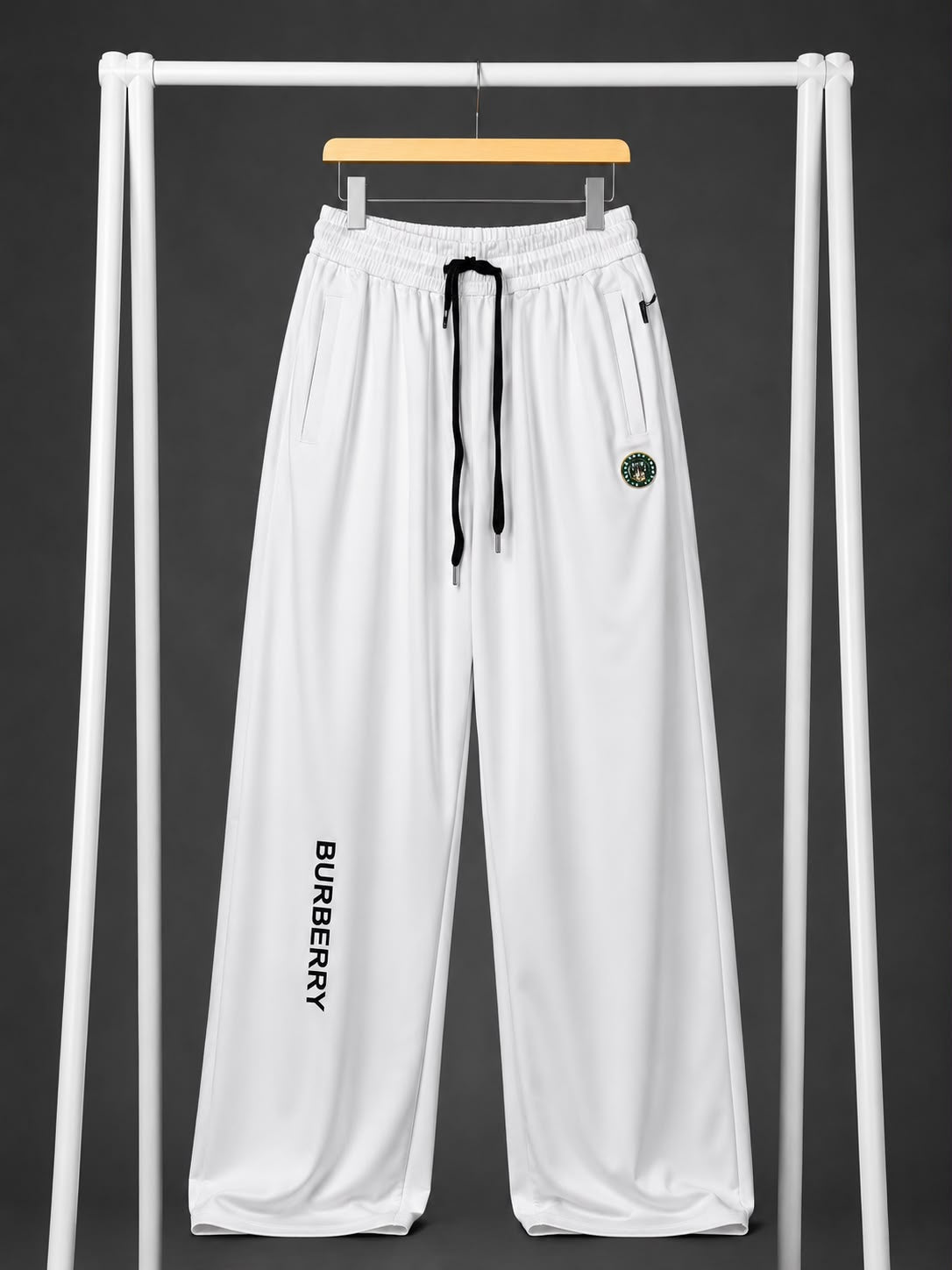 White wide-leg sweatpants hanging on a wooden hanger from a white clothing rack against a dark gray background. The pants feature a black drawstring, a vertical black 'BURBERRY' logo printed on the lower left leg, and a small circular green and gold crest logo on the upper right thigh.