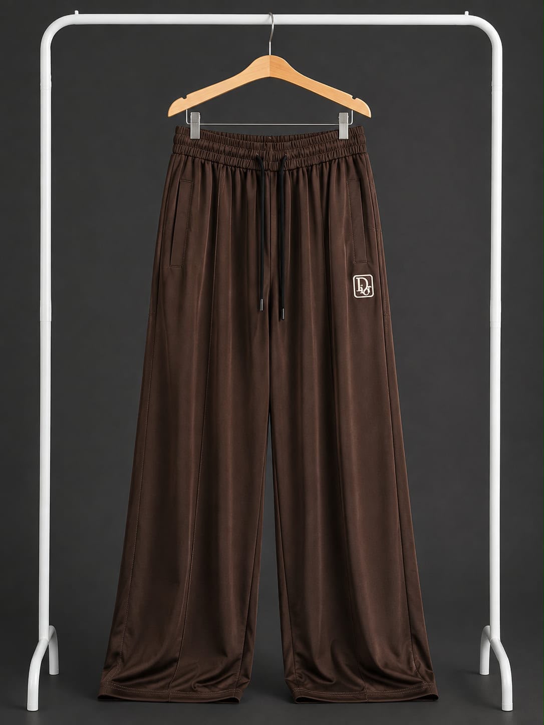 A pair of wide-leg brown pants with an elastic drawstring waist and side pocket, hanging on a wooden hanger from a white clothing rack against a dark gray background.